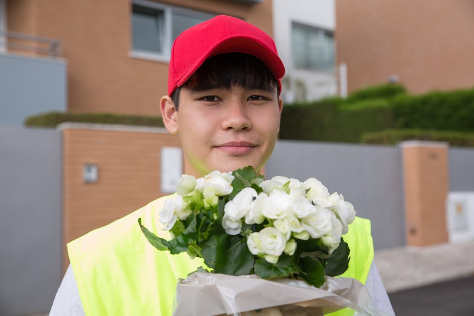 A young male delivery person with a friendly expression, wearing a red cap and high-visibility yellow vest, holds a bouquet of white roses with green leaves wrapped in white paper. He stands outdoors