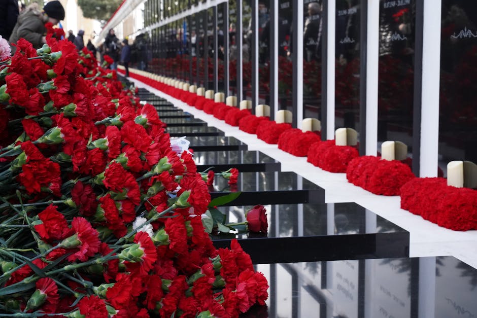 A row of vibrant red carnations and roses arranged as floral tributes along a memorial fence, with red flower bouquets laid on a black polished stone surface. The carnations are densely packed, with s