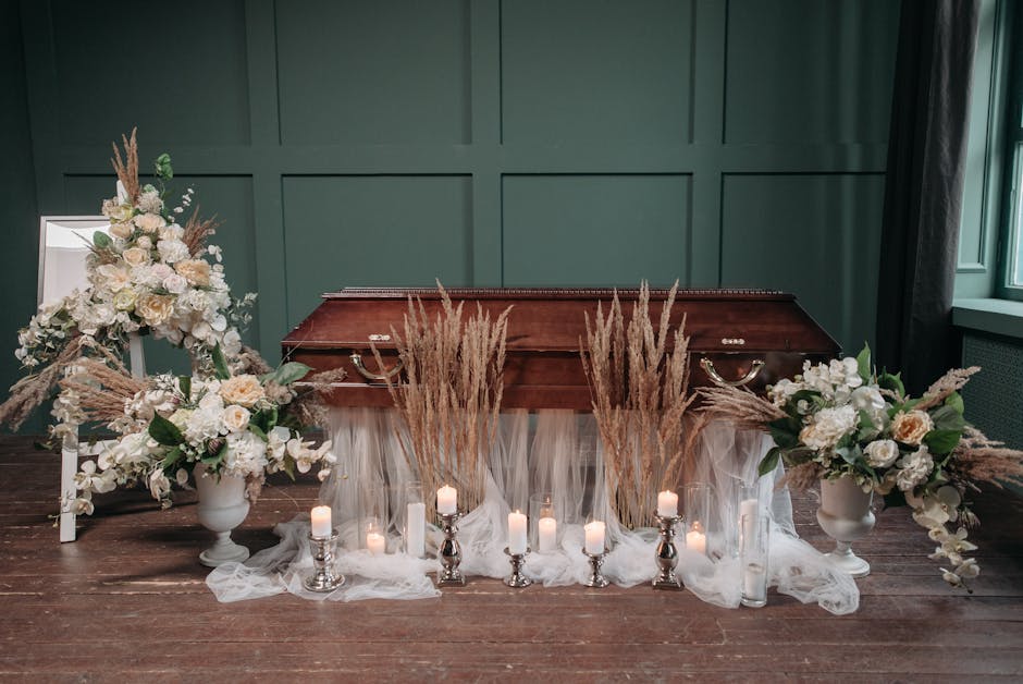 A floral arrangement set in front of a vintage wooden casket, featuring white roses, hydrangeas, and cream-colored carnations, accented with tall beige pampas grass. The arrangement is displayed in wh