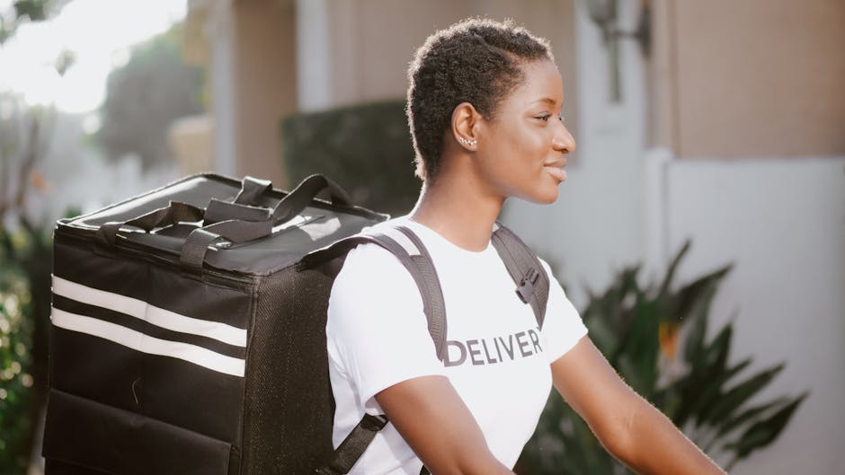 A smiling young woman with short, curly hair, wearing a white T-shirt with the word 'DELIVER' printed on it, is outdoors in natural light. She carries a large black delivery bag with white stripes ove