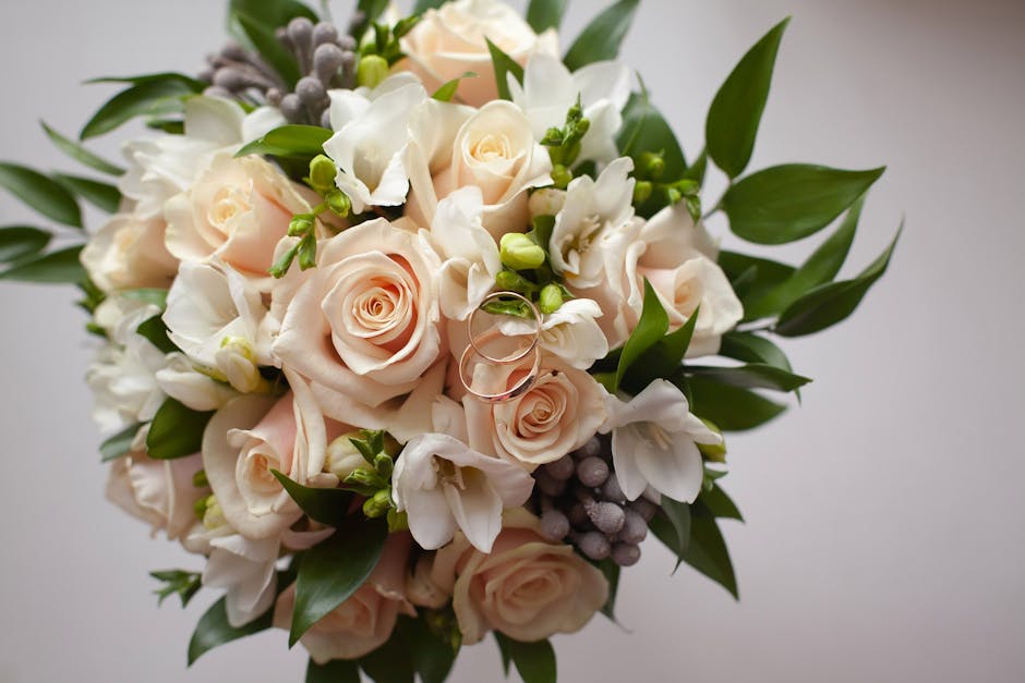 A close-up of a wedding bouquet featuring soft blush pink roses and delicate white freesias intertwined with green leaves and small purple berries. The flowers are fresh, with smooth petals and sturdy