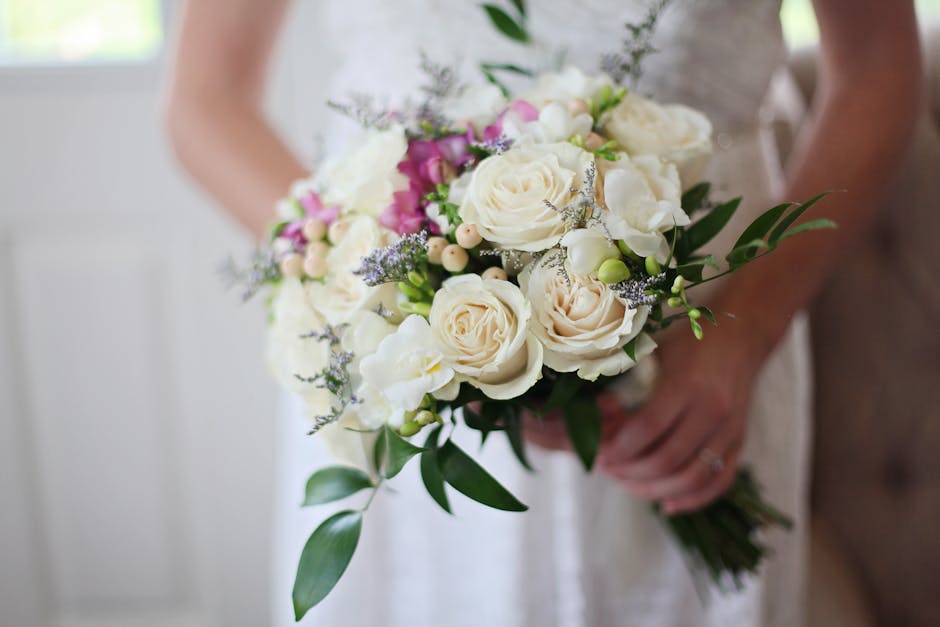 A floral bouquet held by a person, featuring a mix of white and blush roses, small purple flowers, and green foliage. The arrangement is fresh and lush, with softly textured petals and vibrant leaves,